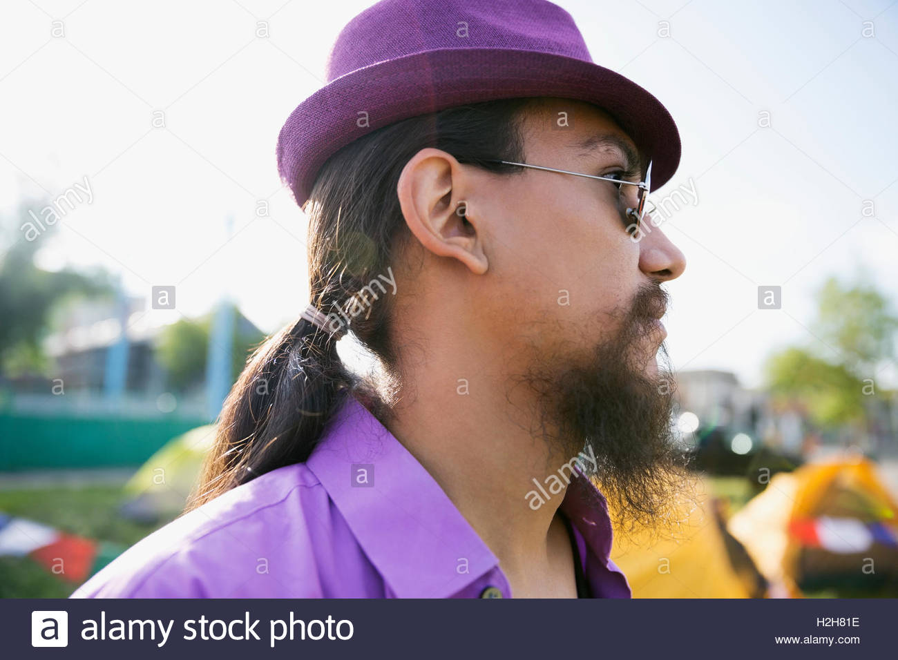 Close up portrait pensive young man with ponytail and beard wearing purple hat looking away Stock Photo