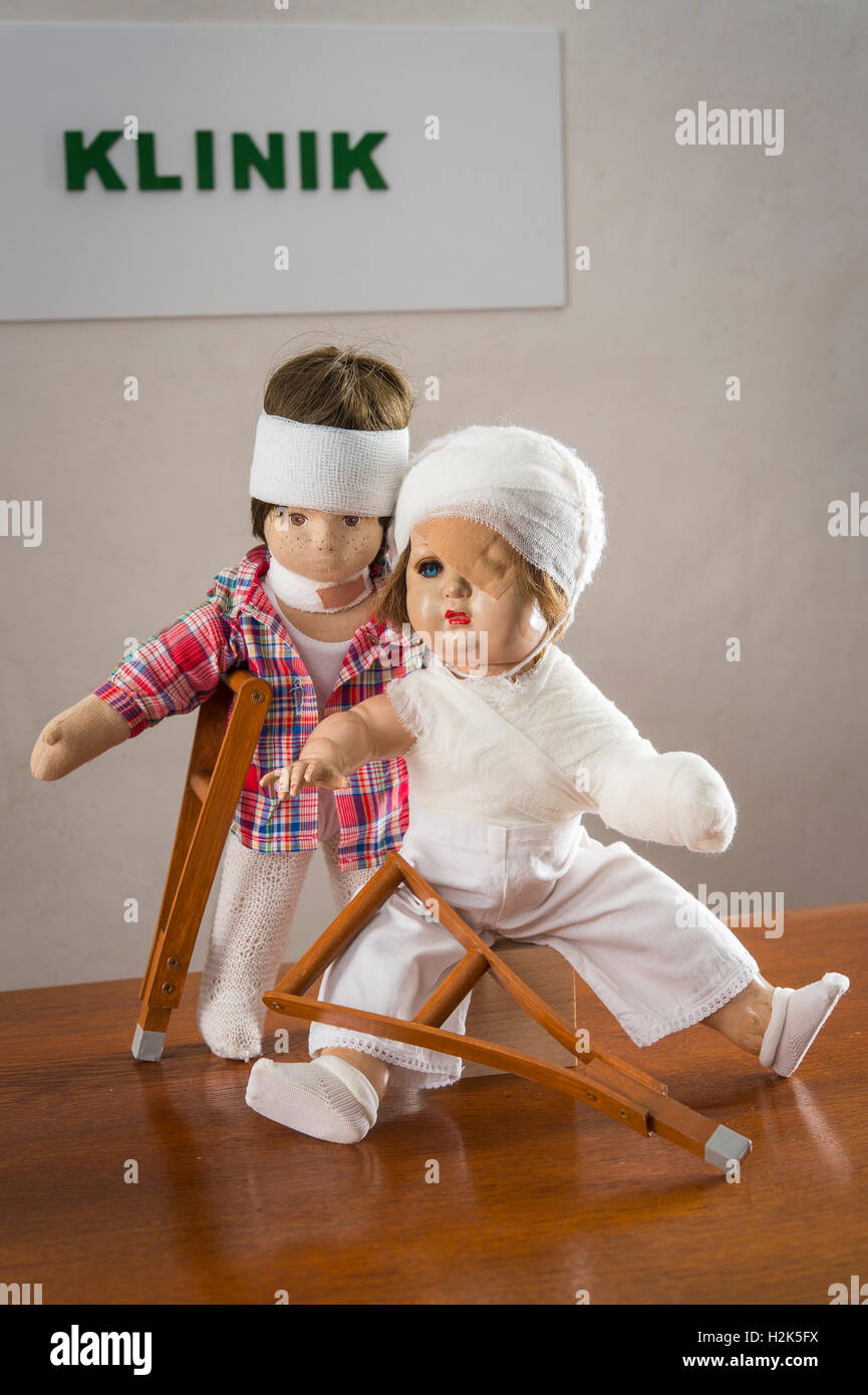 Two dolls, bandages on head, eye patch, arm and shoulder bandage, neck brace, crutches, clinic sign behind Stock Photo