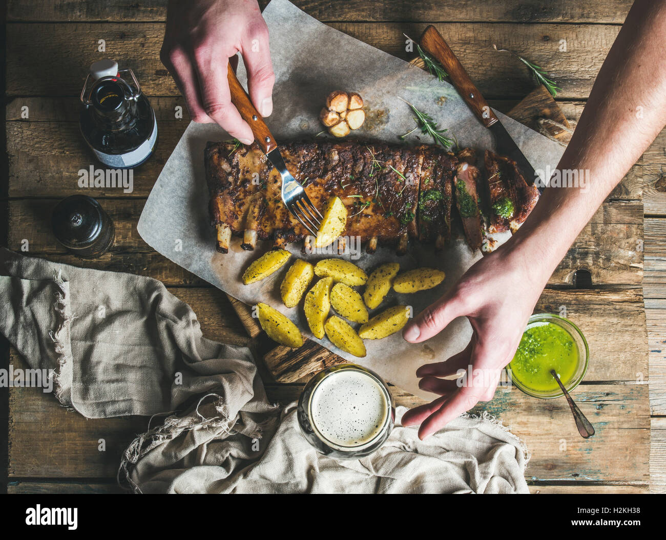 Man eating roasted pork ribs with garlic, rosemary and green herb sauce on rustic wooden table. Man' s hand holding fork with fr Stock Photo