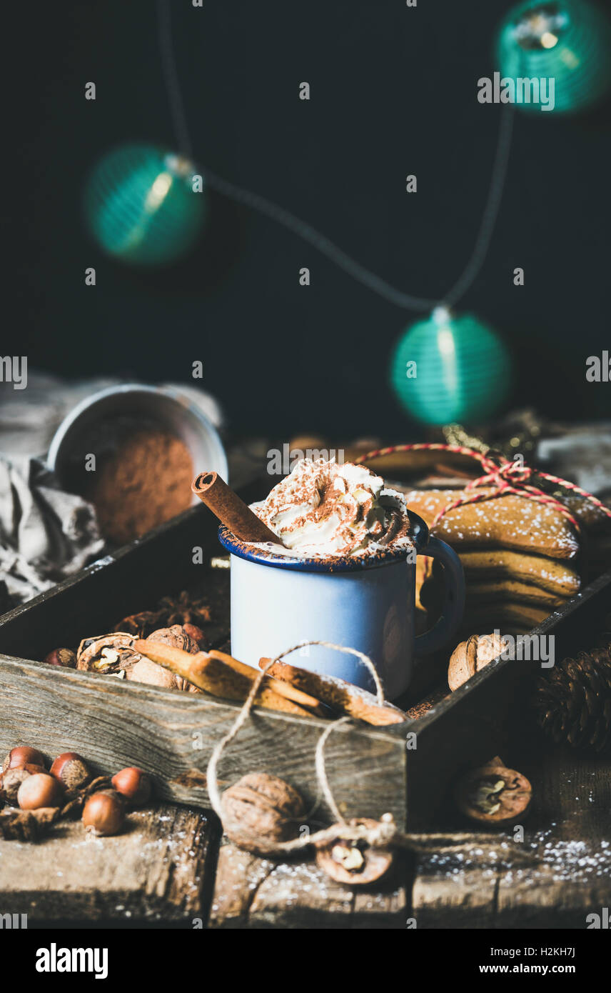 Mug of hot chocolate with whipped cream, cocoa powder, cinnamon, gingerbread cookies, nuts in wooden tray, dark wall with Christ Stock Photo