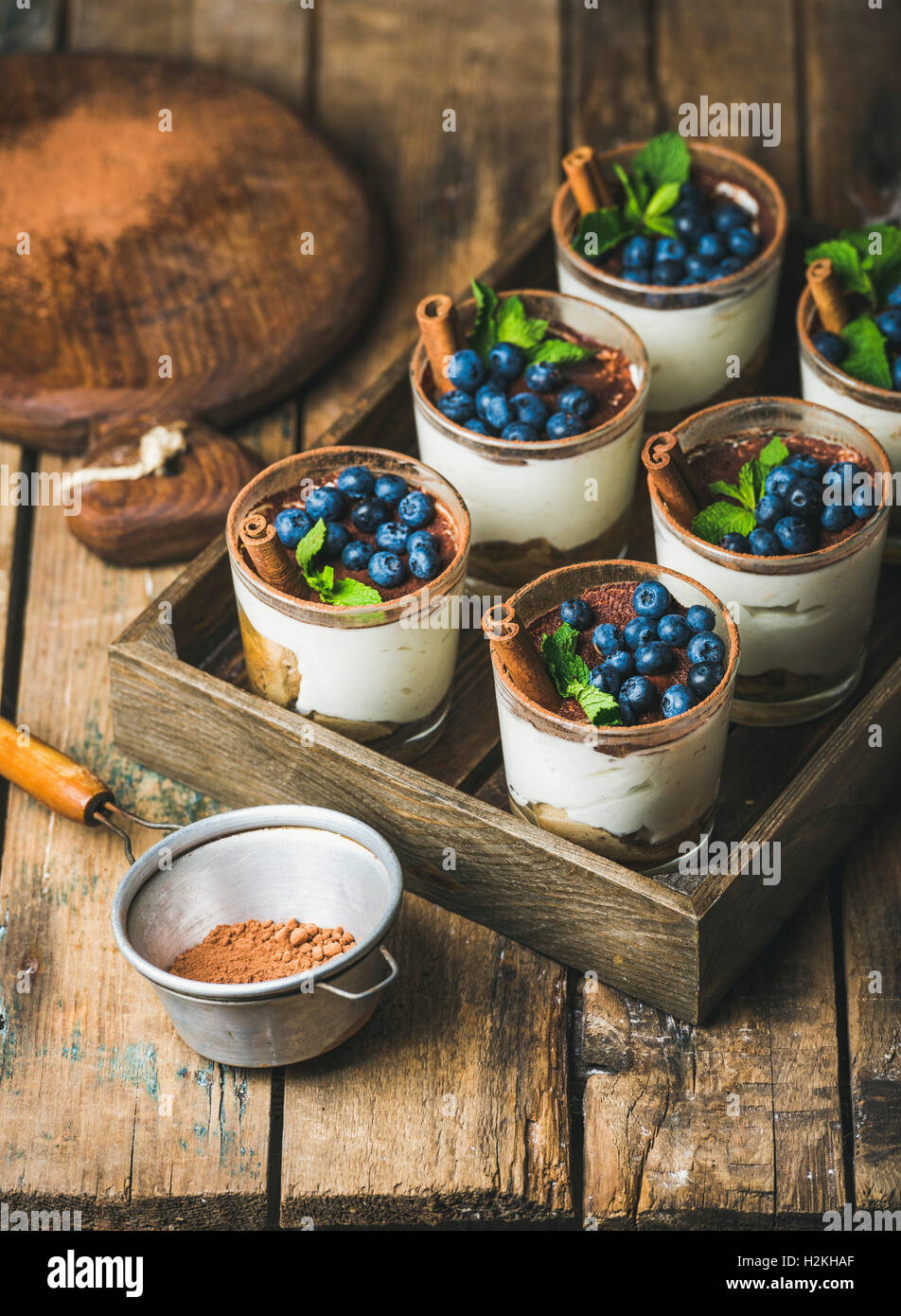 Homemade Tiramisu dessert in glasses with cinnamon, mint and fresh blueberries in wooden tray and sieve with cocoa powder over r Stock Photo