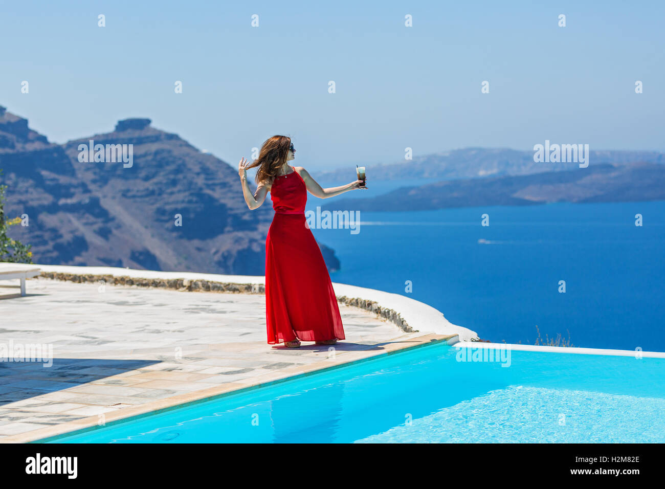 Woman in red dress standing on the edge of the pool and looking out to the sea Stock Photo