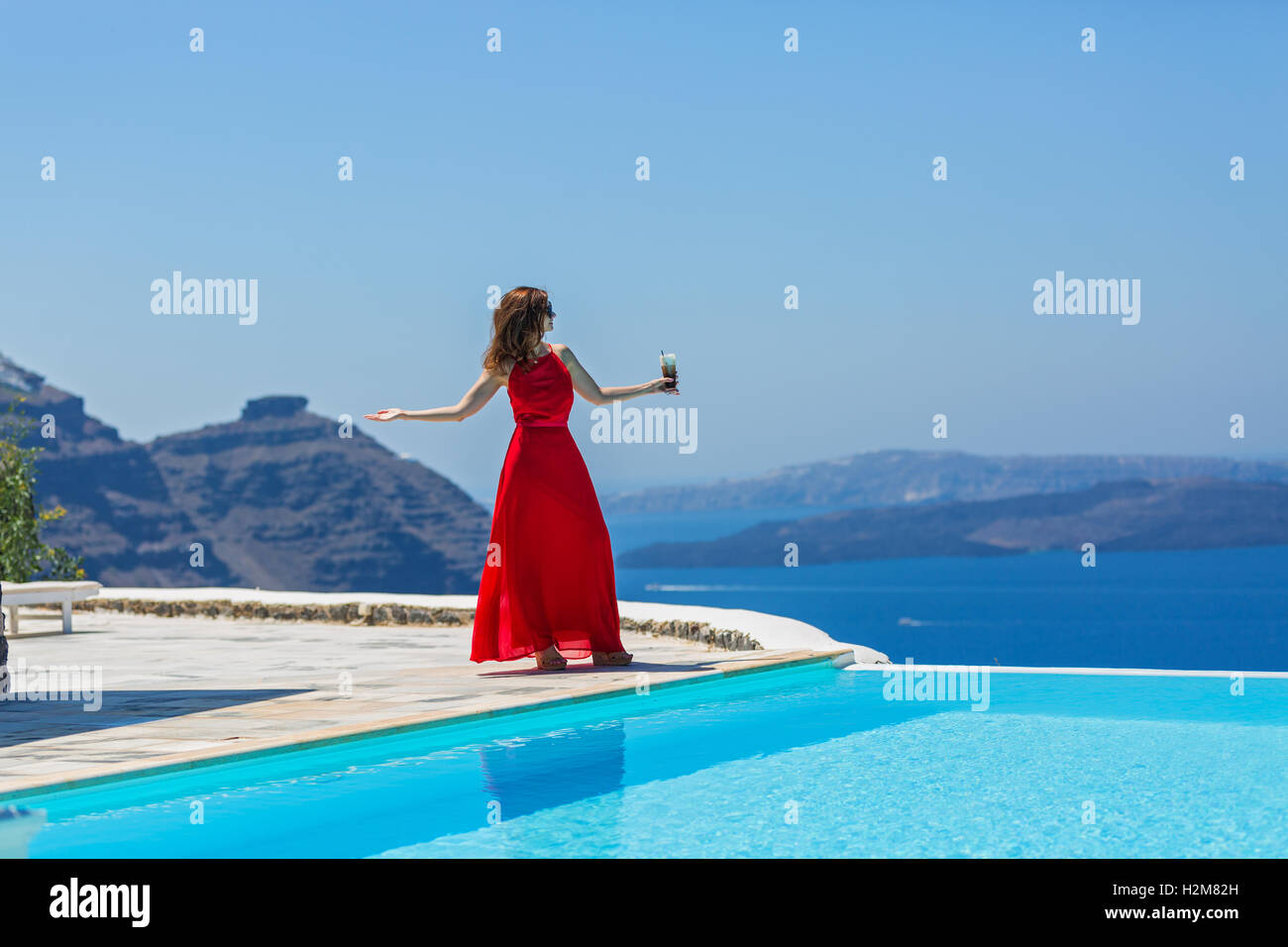 Woman in red dress standing on the edge of the pool and looking out to the sea Stock Photo