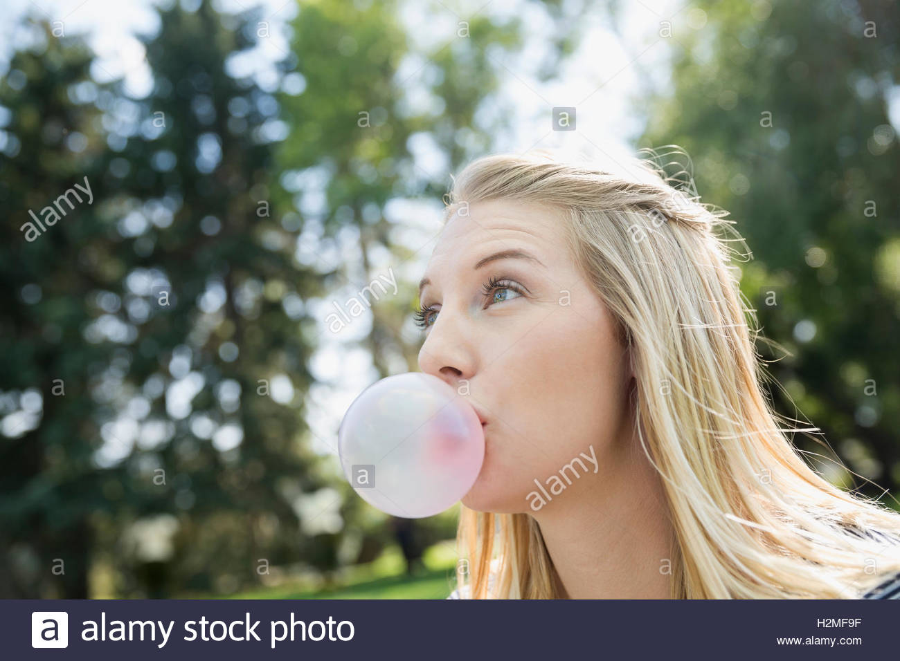 Wide-eyed blonde woman blowing bubble with bubble gum in park Stock Photo