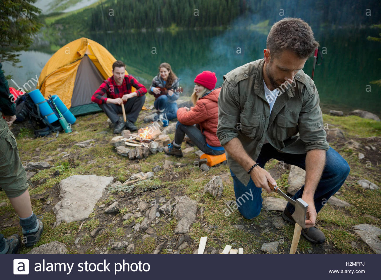 Man splitting wood with ax near campfire at lakeside campsite Stock Photo