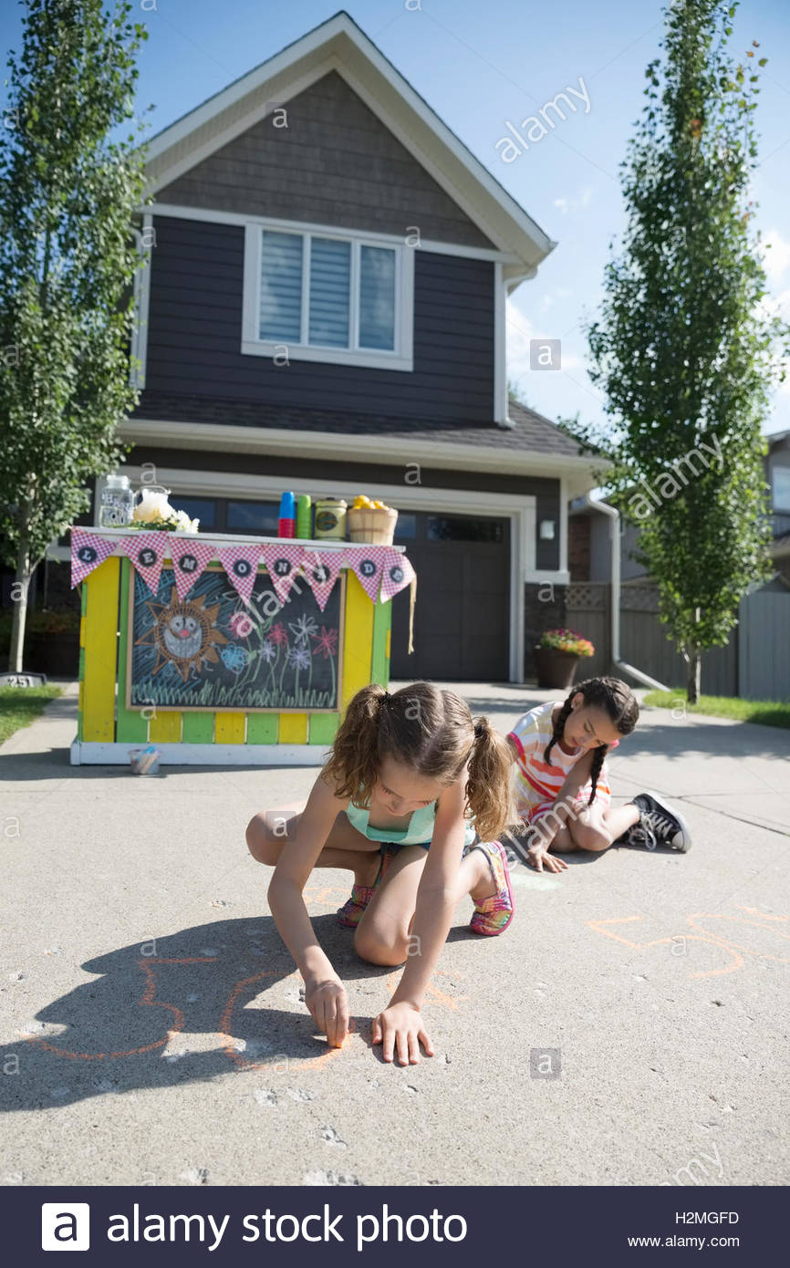 Girls drawing with sidewalk chalk in front of lemonade stand in sunny driveway Stock Photo