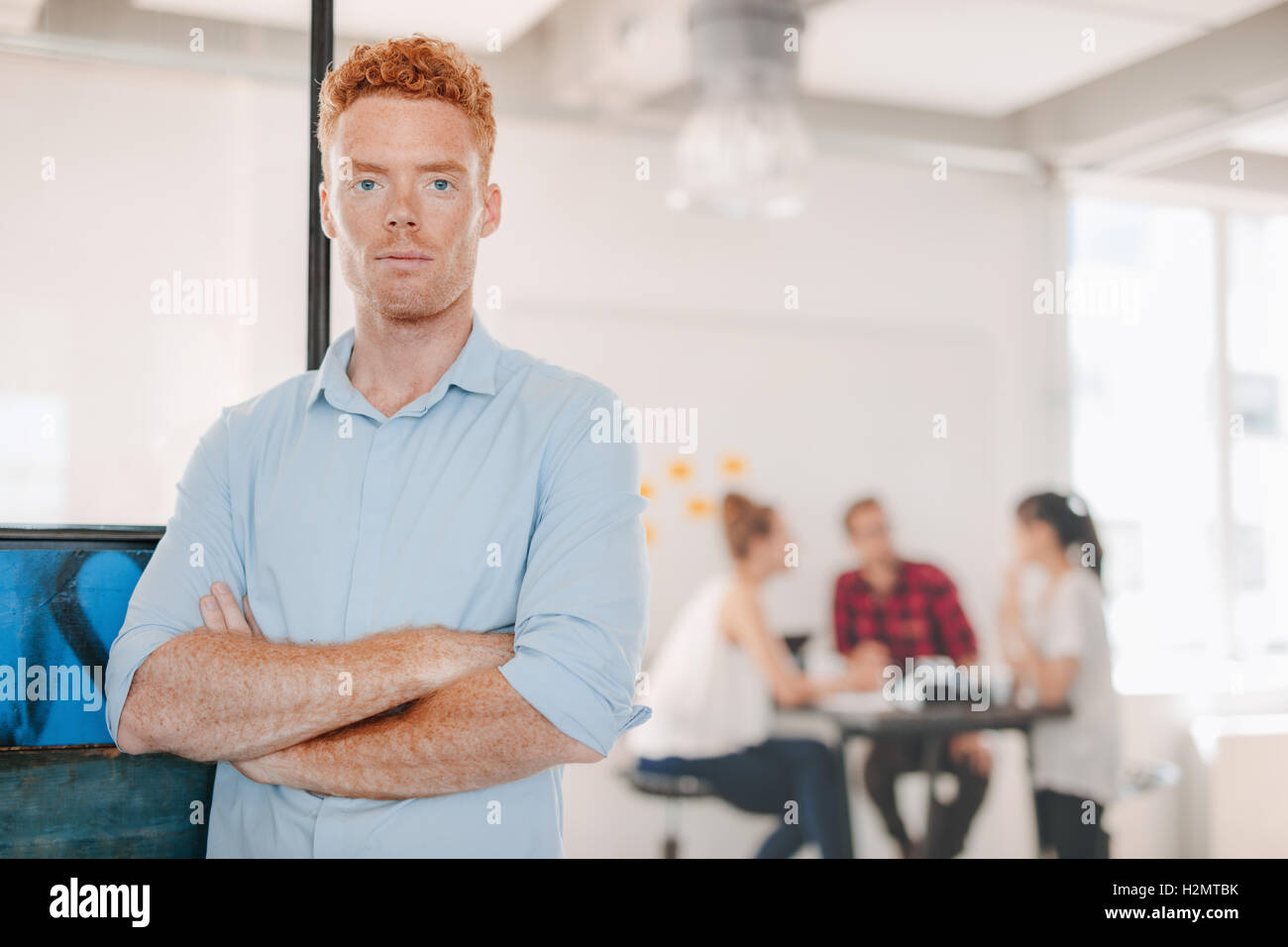 Portrait of confident young businessman standing in office with his arms crossed and colleagues meeting in background. Stock Photo