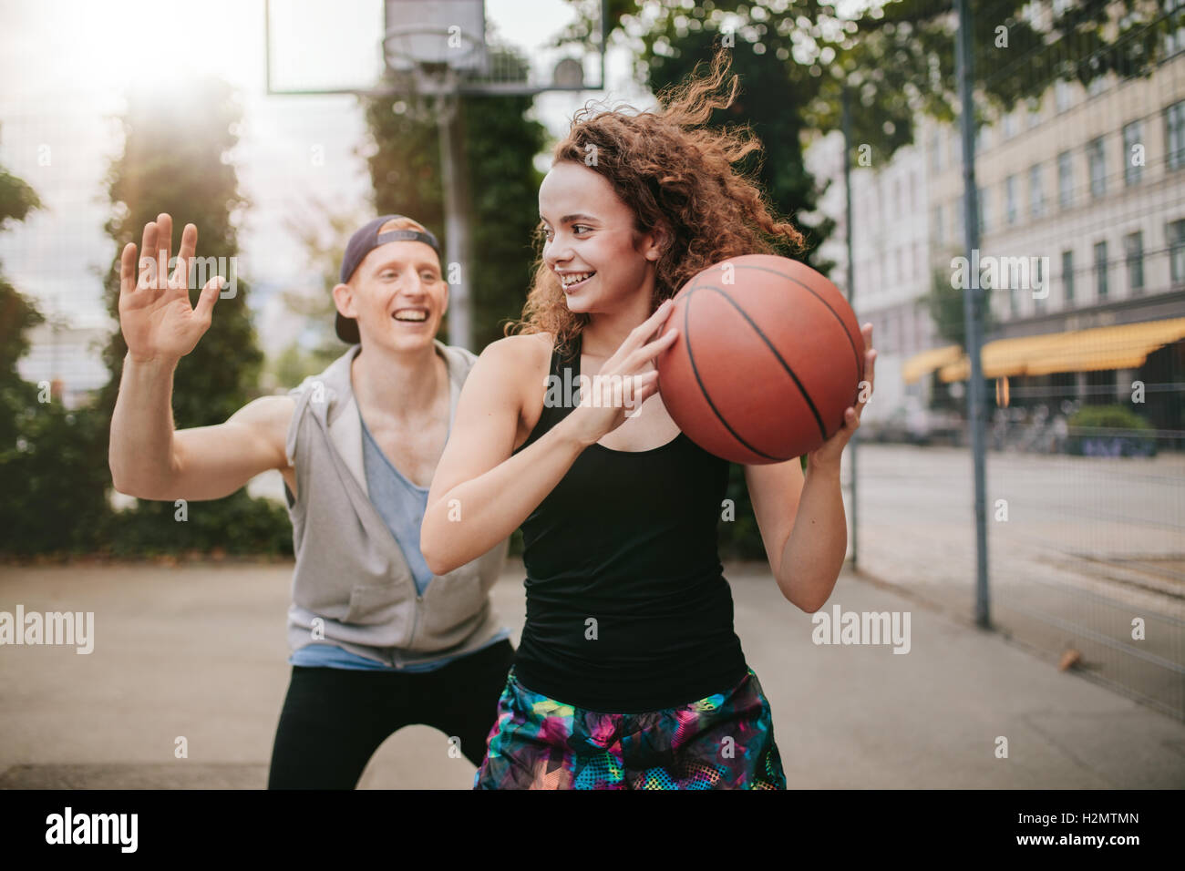 Young girl playing basketball with boy blocking. Teenage friends enjoying a game of streetball on outdoor court. Stock Photo