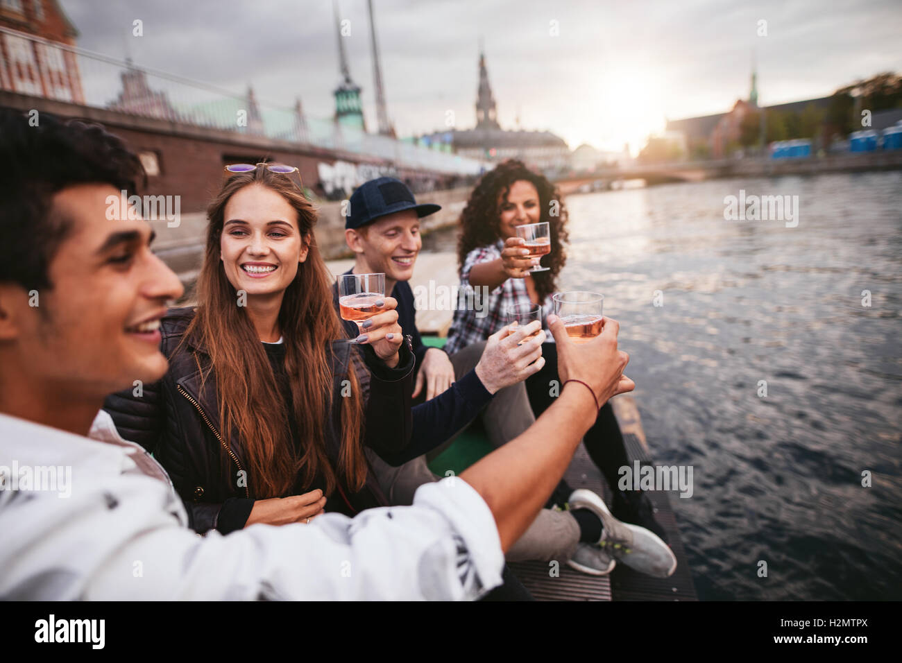 Men and women sitting on jetty and toasting drinks. Group of young friends hanging out by the lake in city. Stock Photo