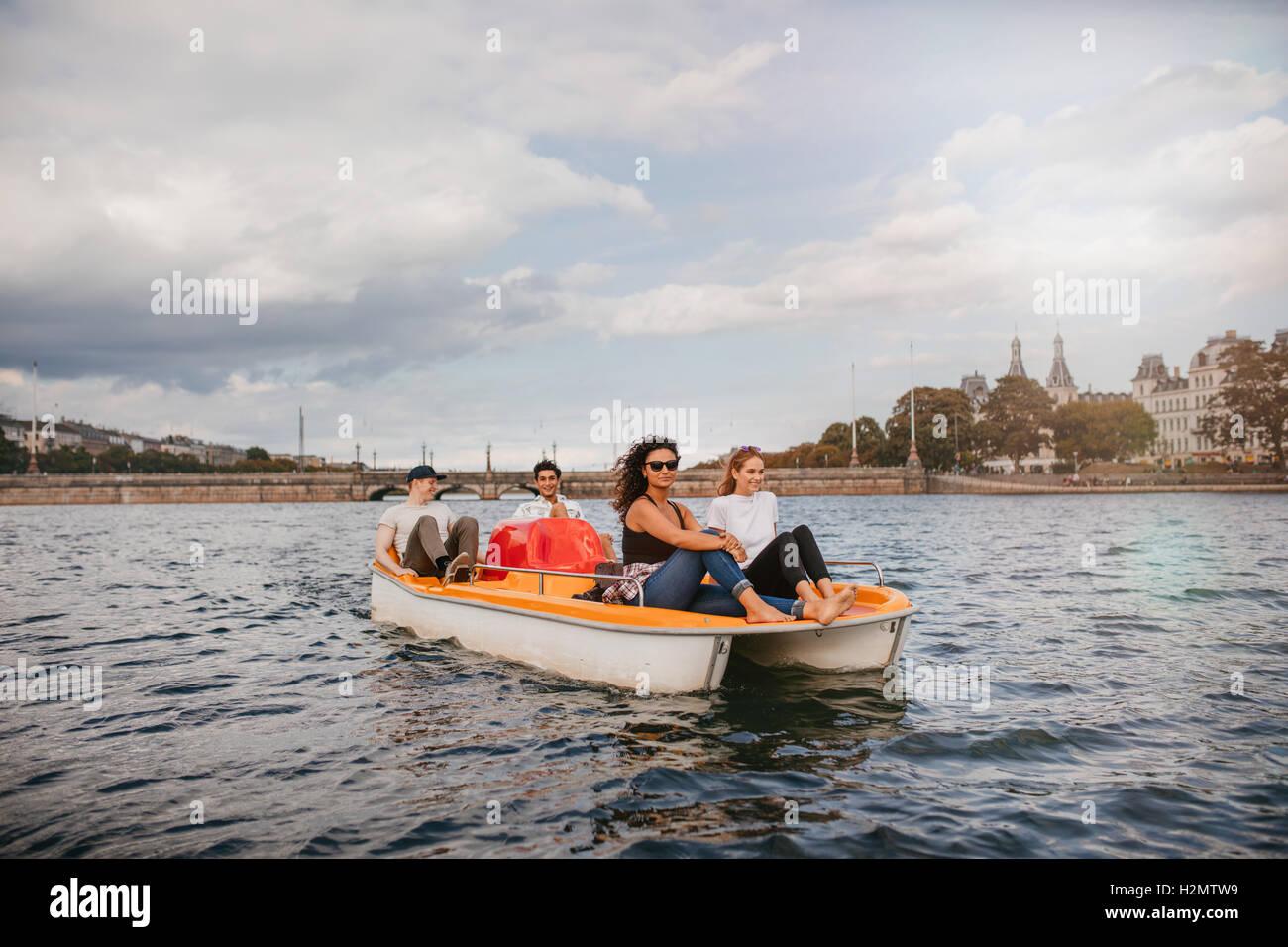 Shot of four young people on pedal boat in lake. Women sitting on front with men at back pedaling the boat. Stock Photo