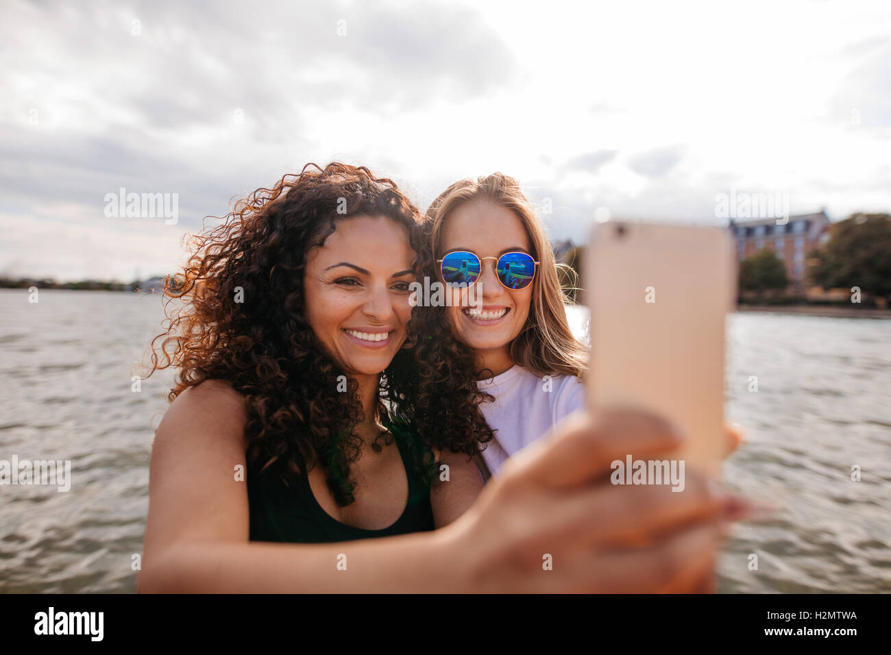 Shot of attractive young women smiling and taking selfie with mobile phone by the lake. Female friends taking self portrait with Stock Photo