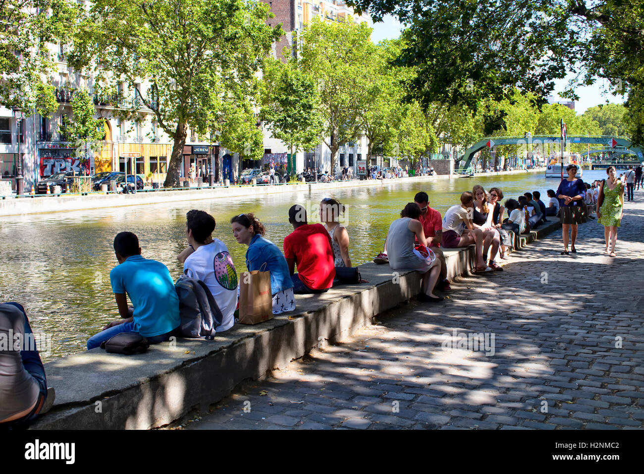 People hang out by Canal Saint Martin under trees in Paris. Shadows create dramatic ambience. Stock Photo
