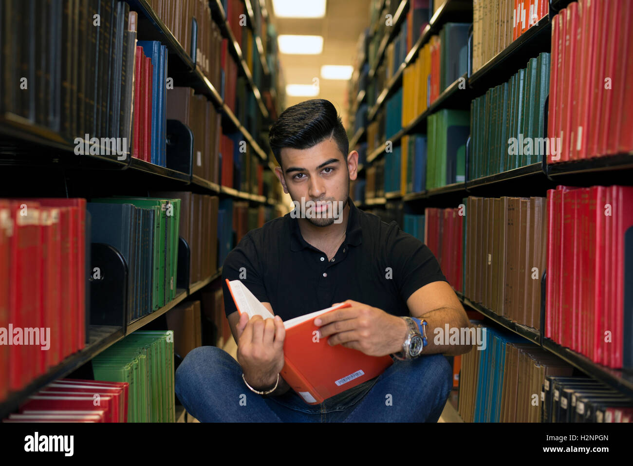 Male college student seen in the library, sitting on the floor between rows of books. He is holding a book, looking at camera. Stock Photo