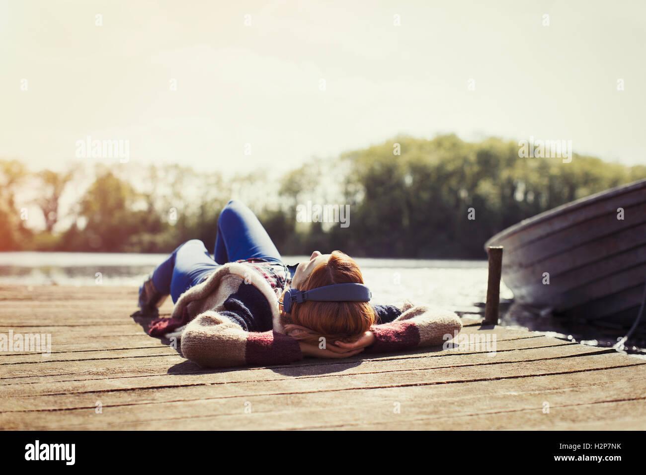 Woman relaxing laying on dock listening to music with headphones at sunny lakeside Stock Photo