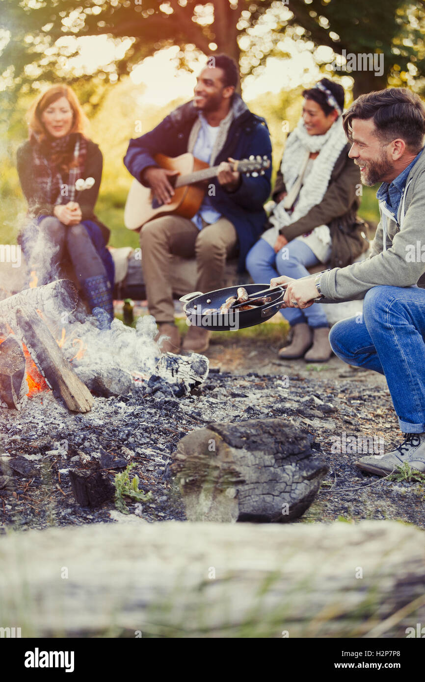 Friends playing guitar and cooking hot dogs at campfire Stock Photo