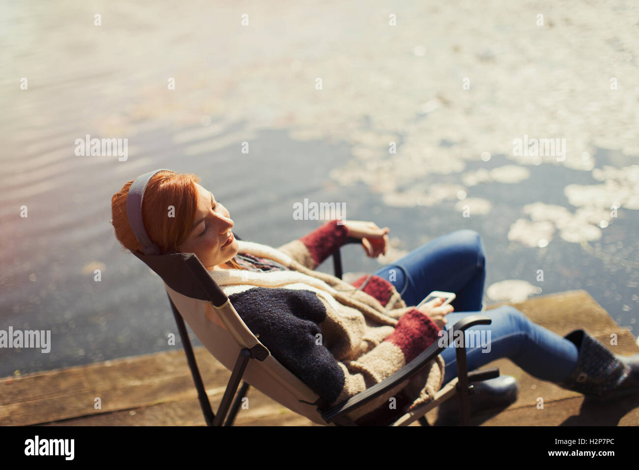 Serene woman relaxing listening to music with headphones and smart phone at sunny lakeside dock Stock Photo