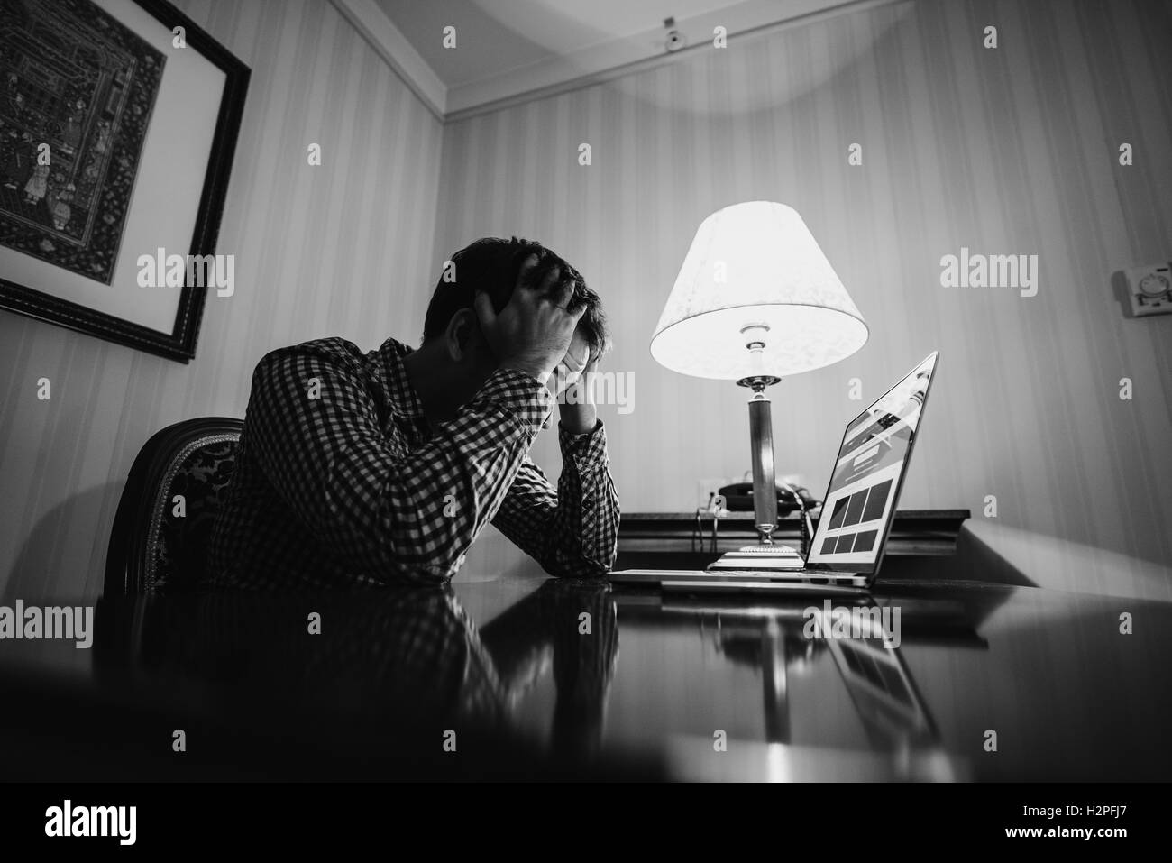 Feeling exhausted. Frustrated young man with hands on his head and keeping eyes closed while sitting at working place. Black white photo. Stock Photo