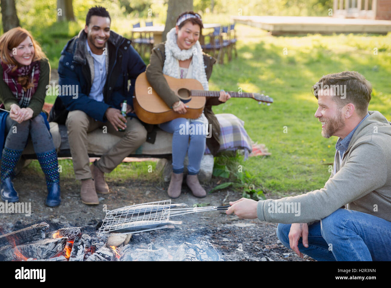 Friends playing guitar and cooking fish in grill basket over campfire Stock Photo