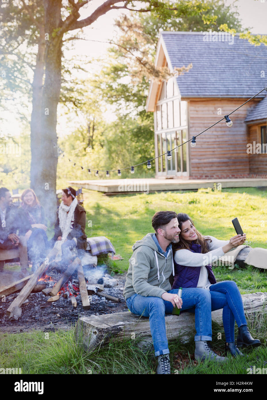Couple taking selfie with camera phone at campfire outside cabin Stock Photo