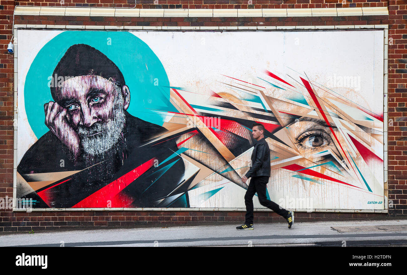 Portrait of old, sad bearded man. Painted faces at the Sun, Spray and Art Festival Art. Sand, Sea & Spray, Blackpool`s Urban Art Festival....Artwork by Otto Schade.in East Topping street, Blackpool, Lancashire, UK Pedestrian walking past street art, graffiti wall painting, and decoration.The Sand, Sea & Spray Urban Art Festival with artwork from Catch 22 and Lowdown designs, stencil based works in Coronation Street, Blackpool, Lancashire, UK Stock Photo