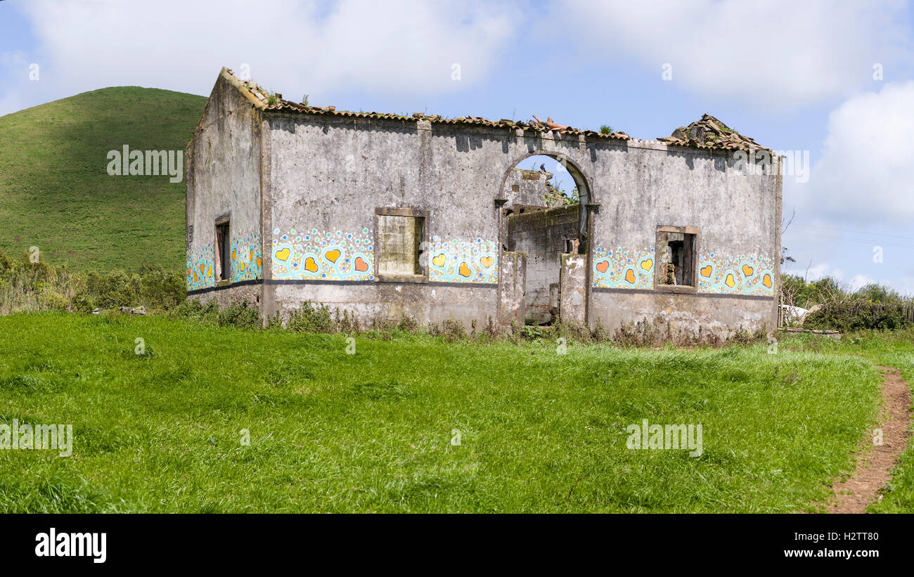 Abandoned Cottage Decorated by Yves Decoster. A rundown, roofless cottage set in a lush green pasture with a hill behind Stock Photo