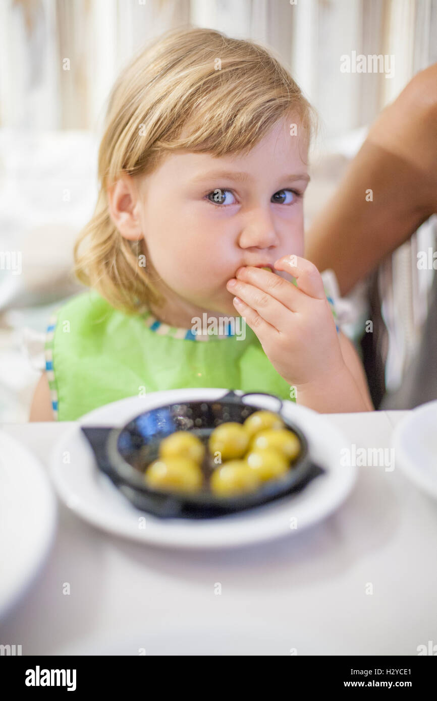 three years old child with green bib looking and eating olives sitting next to woman legs at