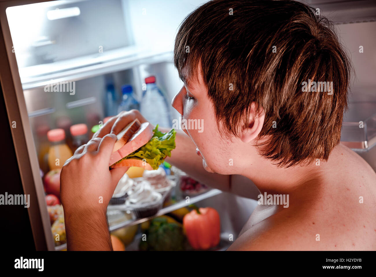 Hungry fat man holding a big sandwich in his hands and standing next to the open fridge. Unhealthy food. Stock Photo