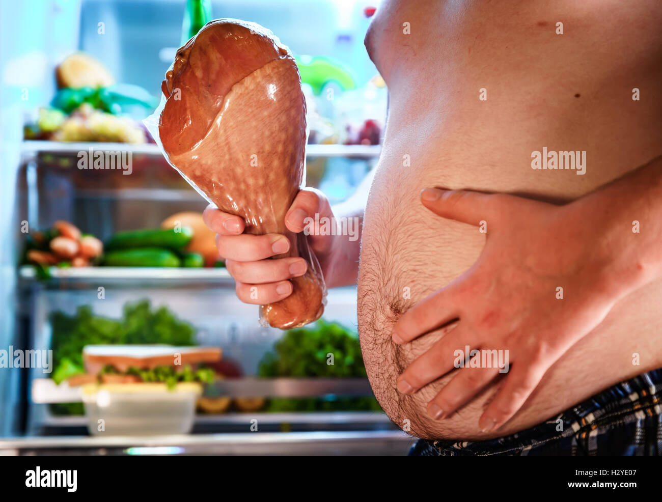 Hungry fat man holding a big turkey leg in his hands and standing next to the open fridge. Unhealthy food. Stock Photo