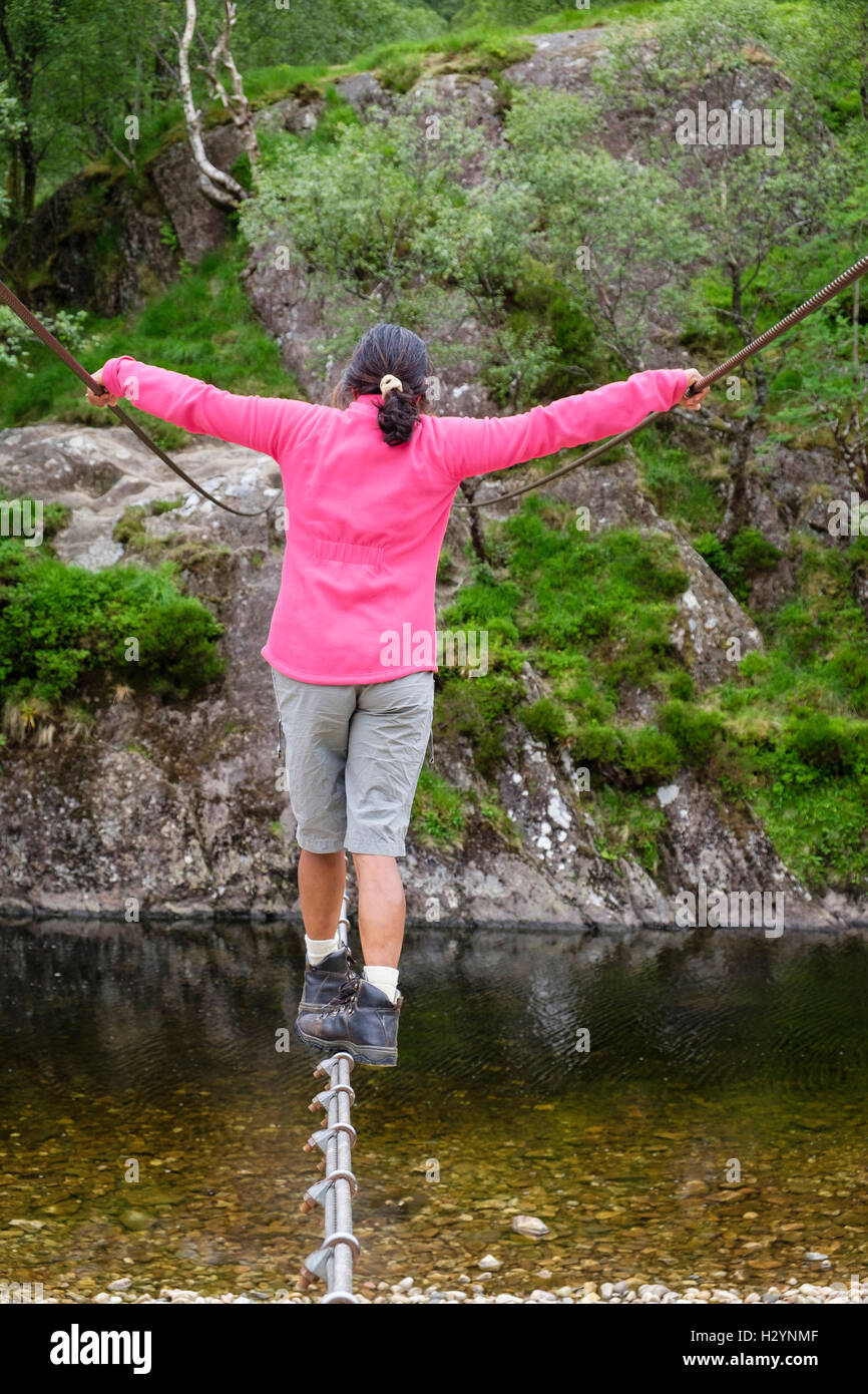 Back view of a woman balancing to walk across Steall wire rope bridge crossing River Nevis. Glen Nevis Fort William Scotland UK Britain Stock Photo