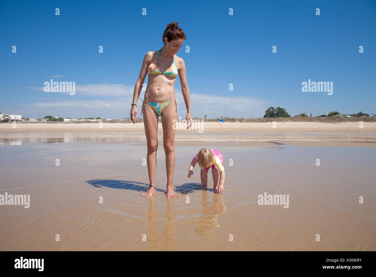 summer family of two years old blonde baby with pink and yellow swimsuit  holding hand with brunette woman mother in bikini stand Stock Photo - Alamy