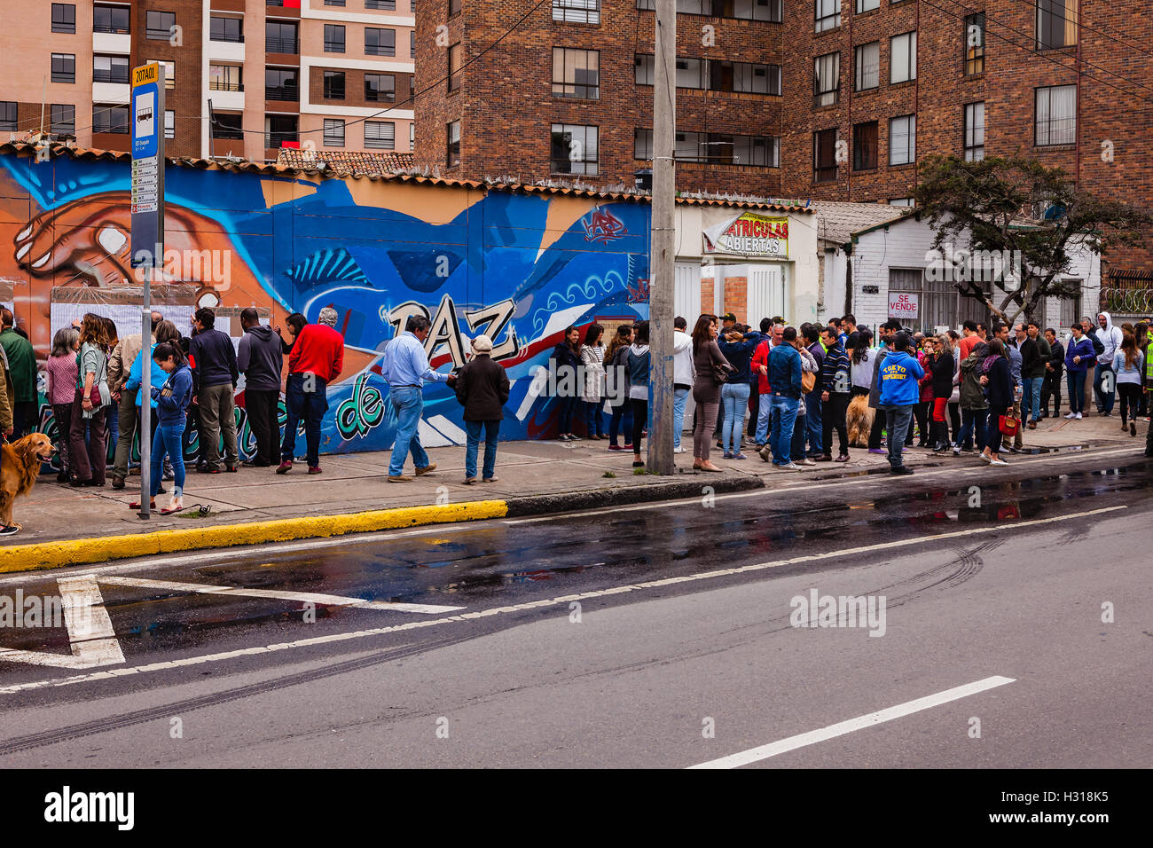 Bogota, Colombia - October 02, 2016: Voters queue up on the right, to enter a polling station on Carrera 9, in the Andean capital city of Bogota, in the South American country of Colombia, to vote on the historic Plebiscite on the peace process with FARC.To the left, behind the people is part of a mural painted in the bright colours of Latin America, dedicated to the Peace Process; the word, Paz, Spanish for peace, is partially visible. Stock Photo