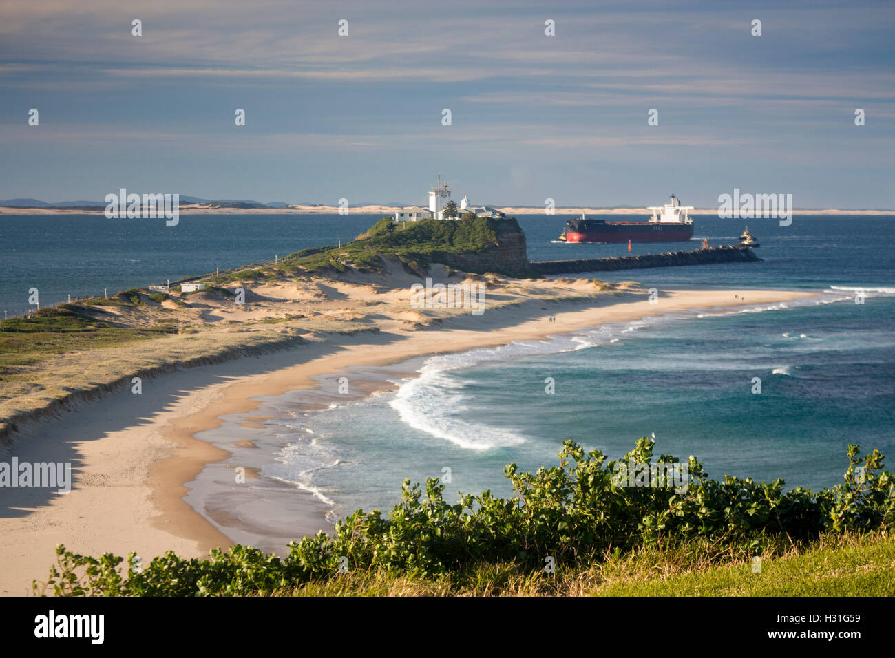 Nobbys Beach and lighthouse with ship being guided by tugs through estuary of Hunter river Newcastle NSW Australia Stock Photo