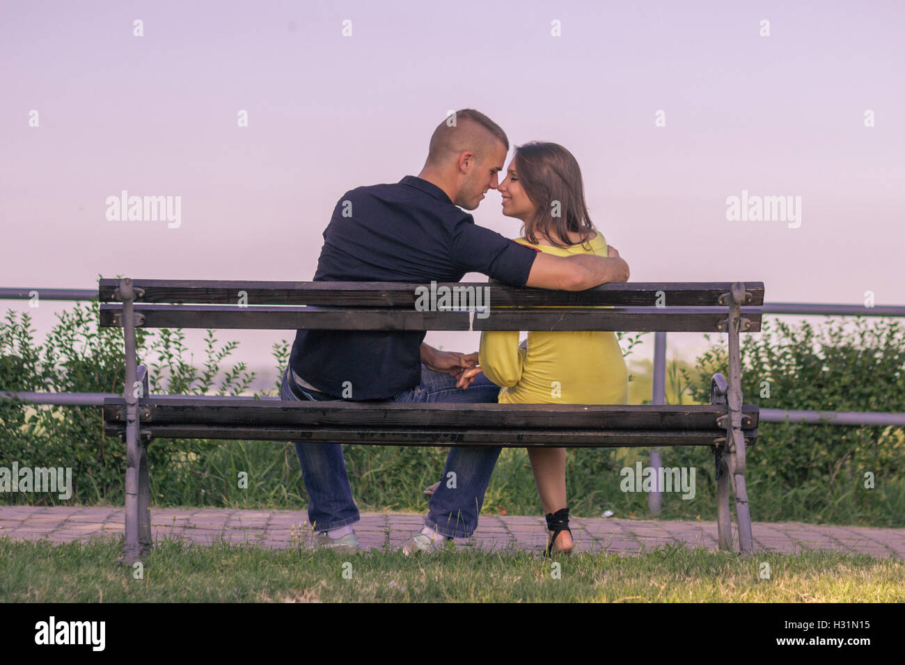 couple 20s sitting bench holding hands touching nose head sky Stock Photo