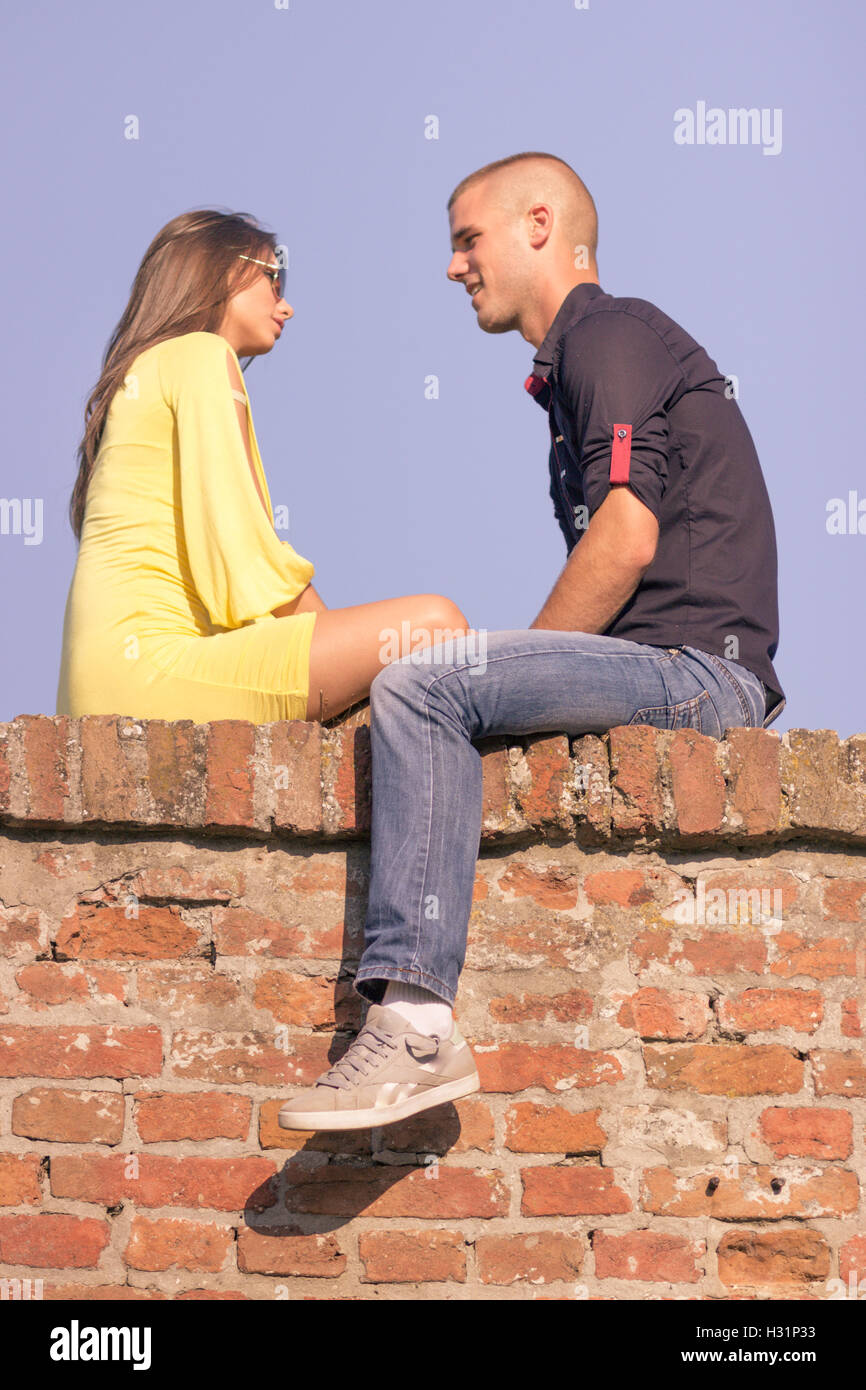 young adult couple 20s sitting brick wall talking sky Stock Photo
