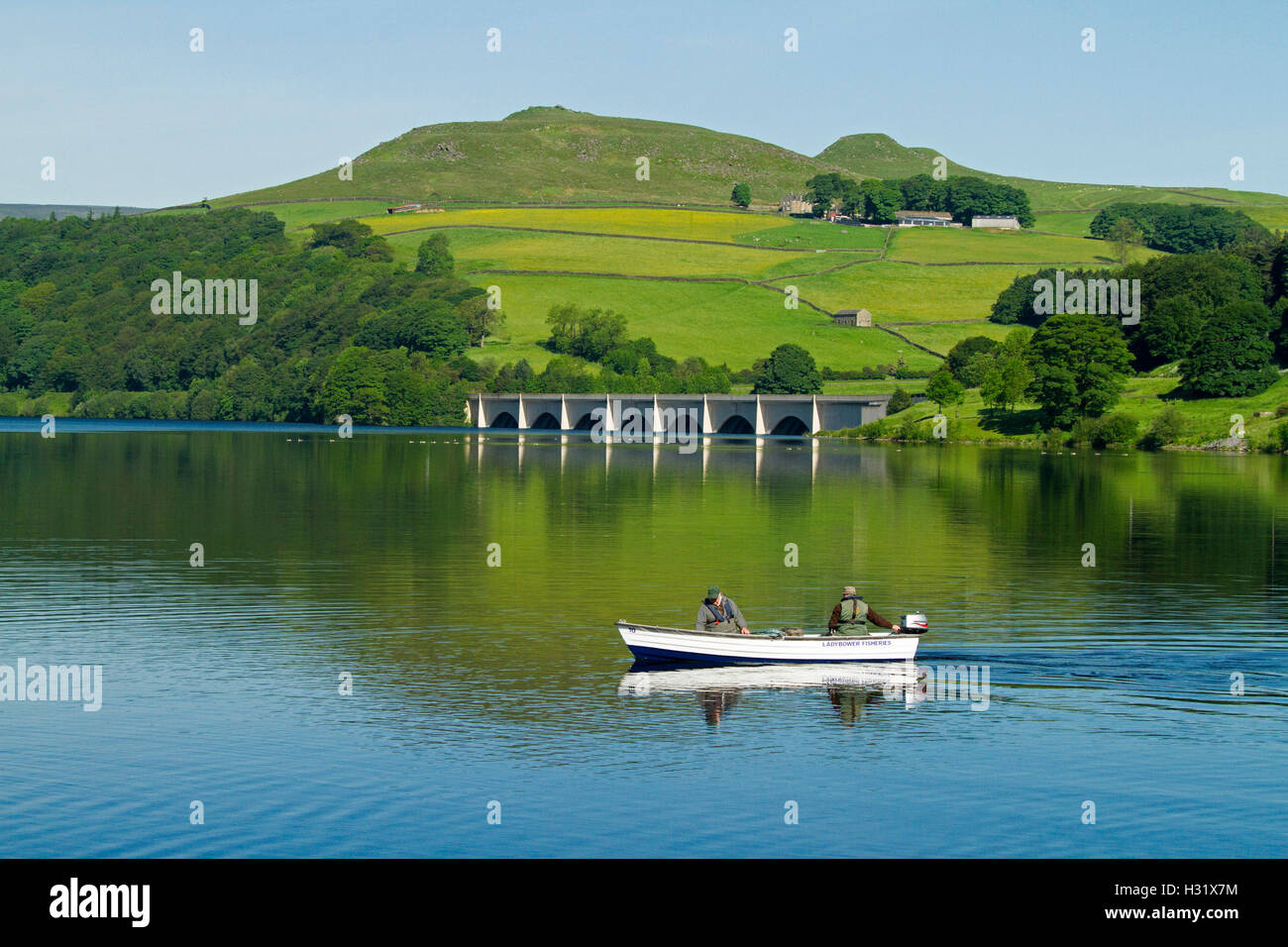 Fishermen in boat on calm blue waters of Ladybower Lake, Ashopton viaduct & farmlands in background under blue sky in England Stock Photo