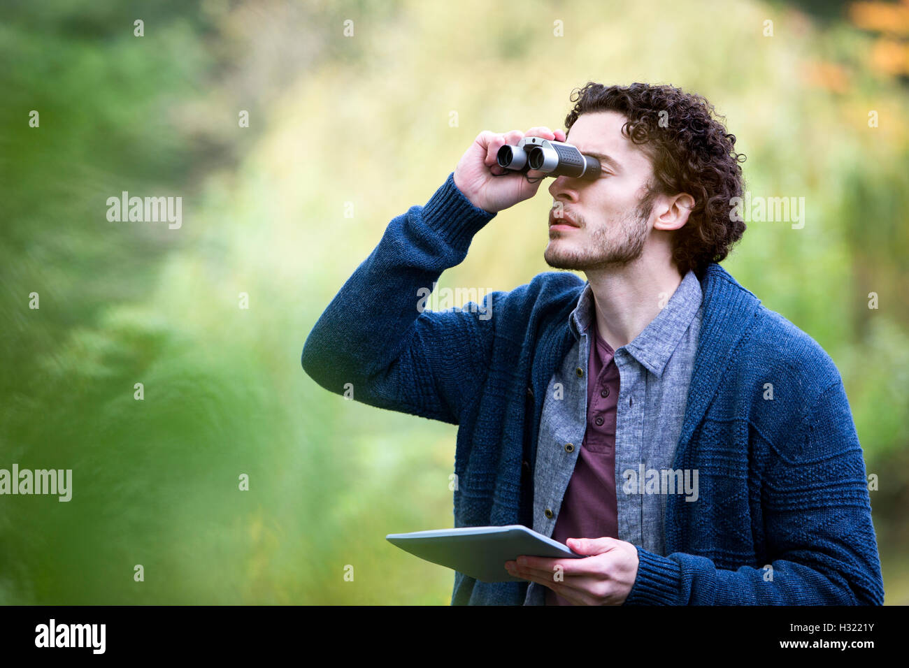 Young man analysing nature with binoculars. He is using a digital tablet to research. Stock Photo