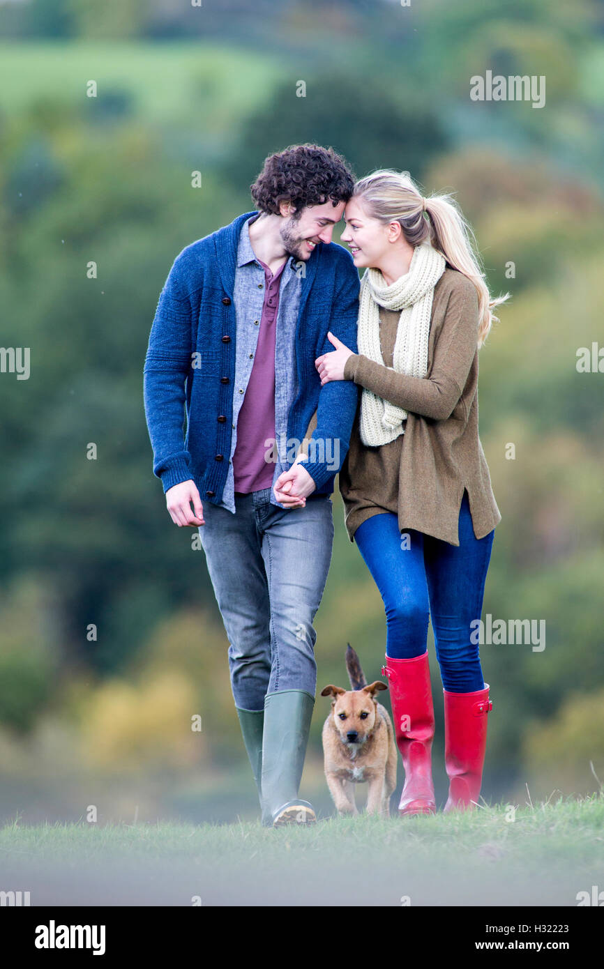 Young couple walking together on a rainy day with their pet dog. They have their heads together and are holding hands. Stock Photo
