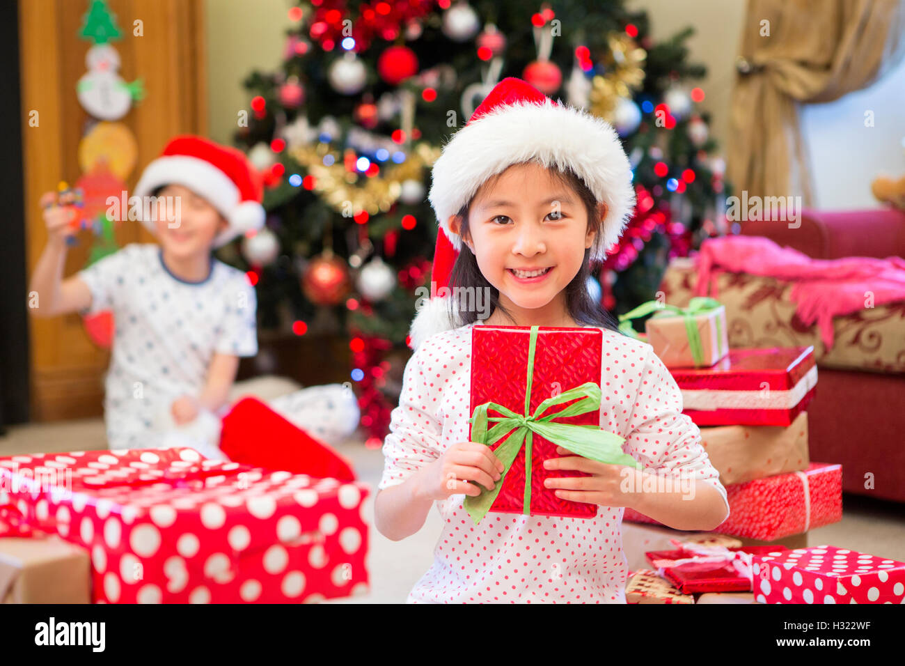 Happy Chinese girl sitting on the floor in her living room at Christmas time. She is surrounded by presents, one of which she is Stock Photo