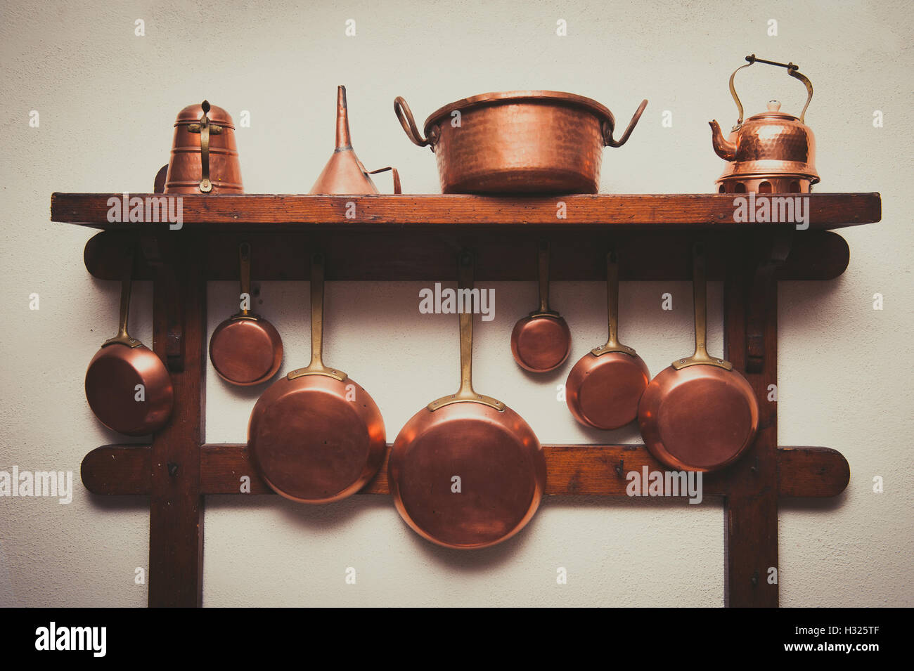 Different kind of vintage copper cookware, pans, coffee pot and funnel hanging on wooden shelf in kitchen with white rough wall Stock Photo