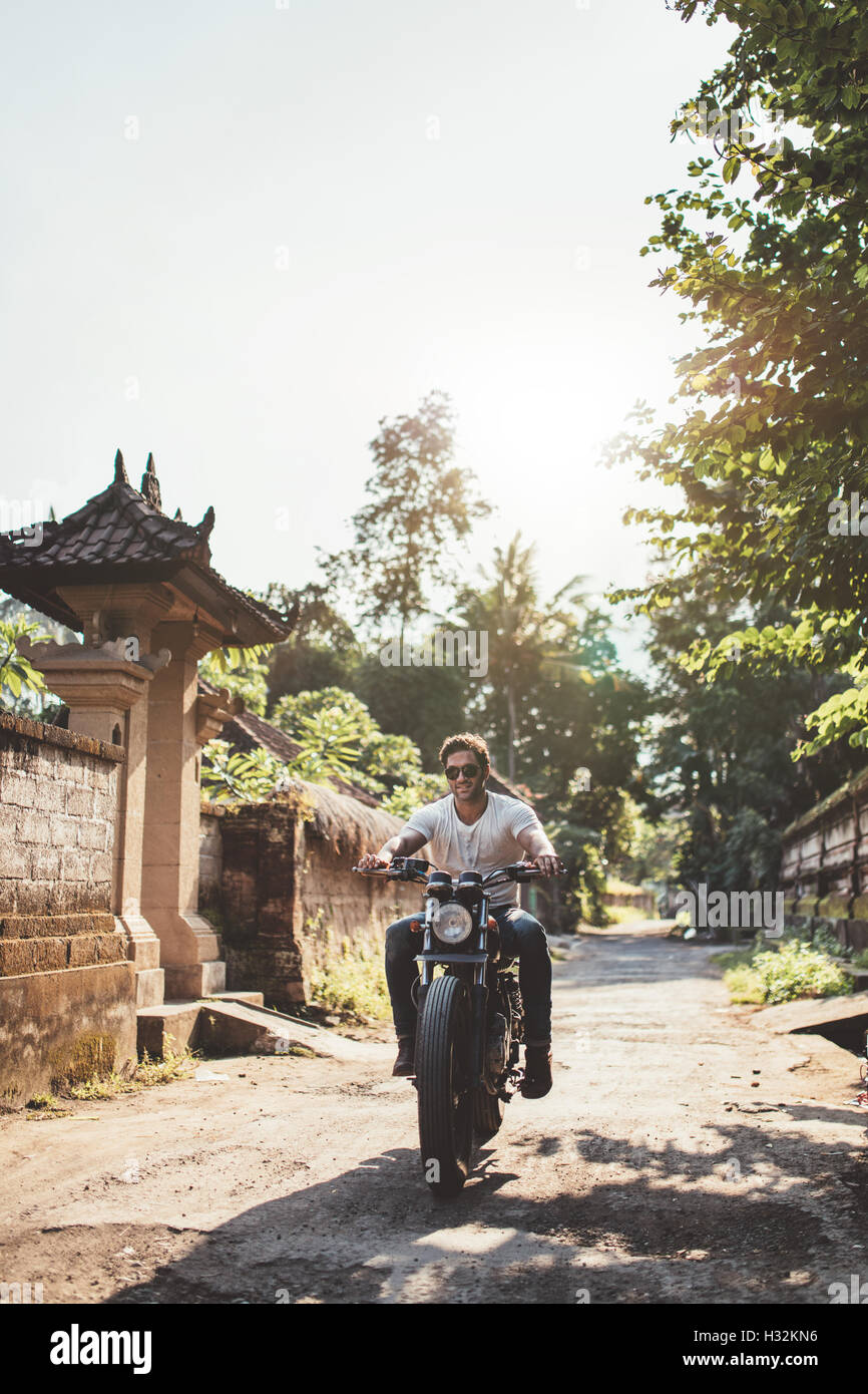 Vertical shot of young man riding motorcycle with her girlfriend through a village road. Couple out on a motorbike ride. Stock Photo