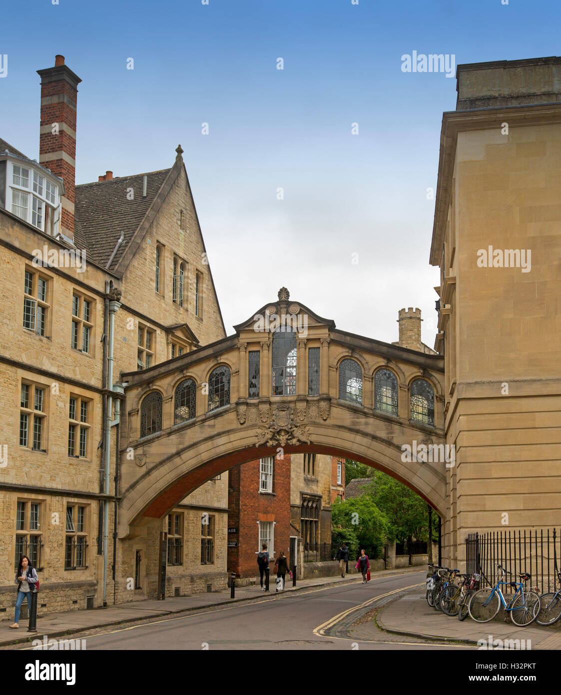 Ornate Bridge of Sighs in Oxford, with bicycles leaning against wall of adjacent  building, pedestrians passing by & blue sky, England Stock Photo