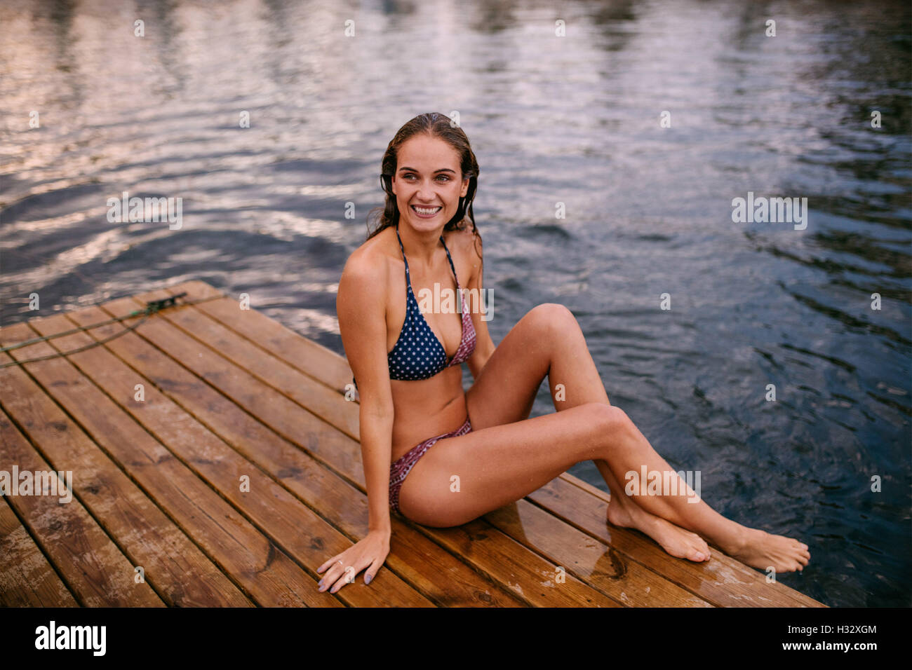 Shot of beautiful young woman in bikini sitting on jetty by the lake. Caucasian teenage girl relaxing on pier looking away and s Stock Photo