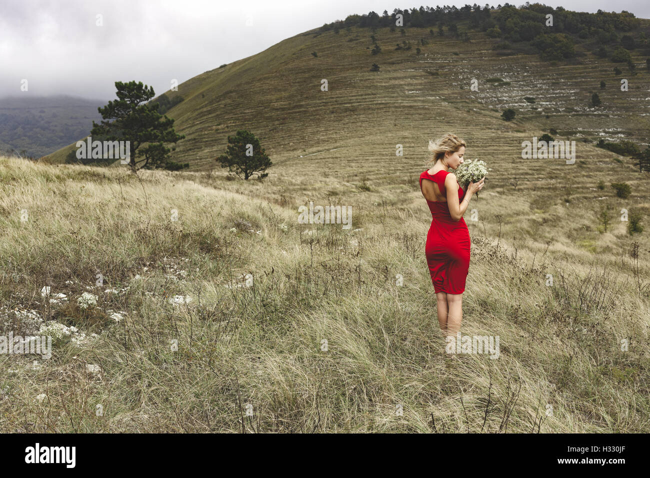 Woman in red dress holding a field flowers while standing in a meadow against terraced slope on a cloudy windy day Stock Photo