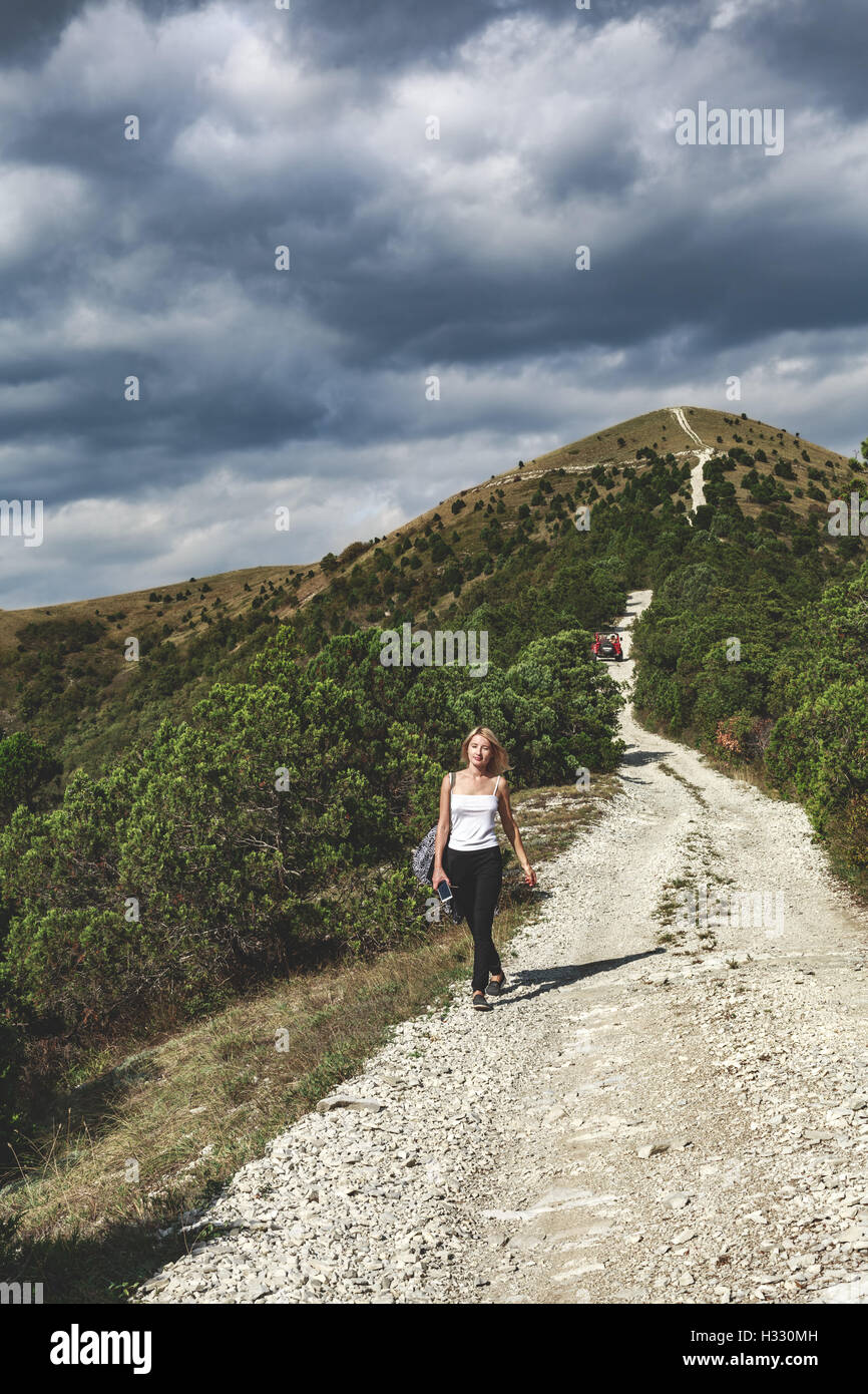 Woman walking down along a mountain road on a cloudly autumn day. Hiking, tourism, adventure concept Stock Photo