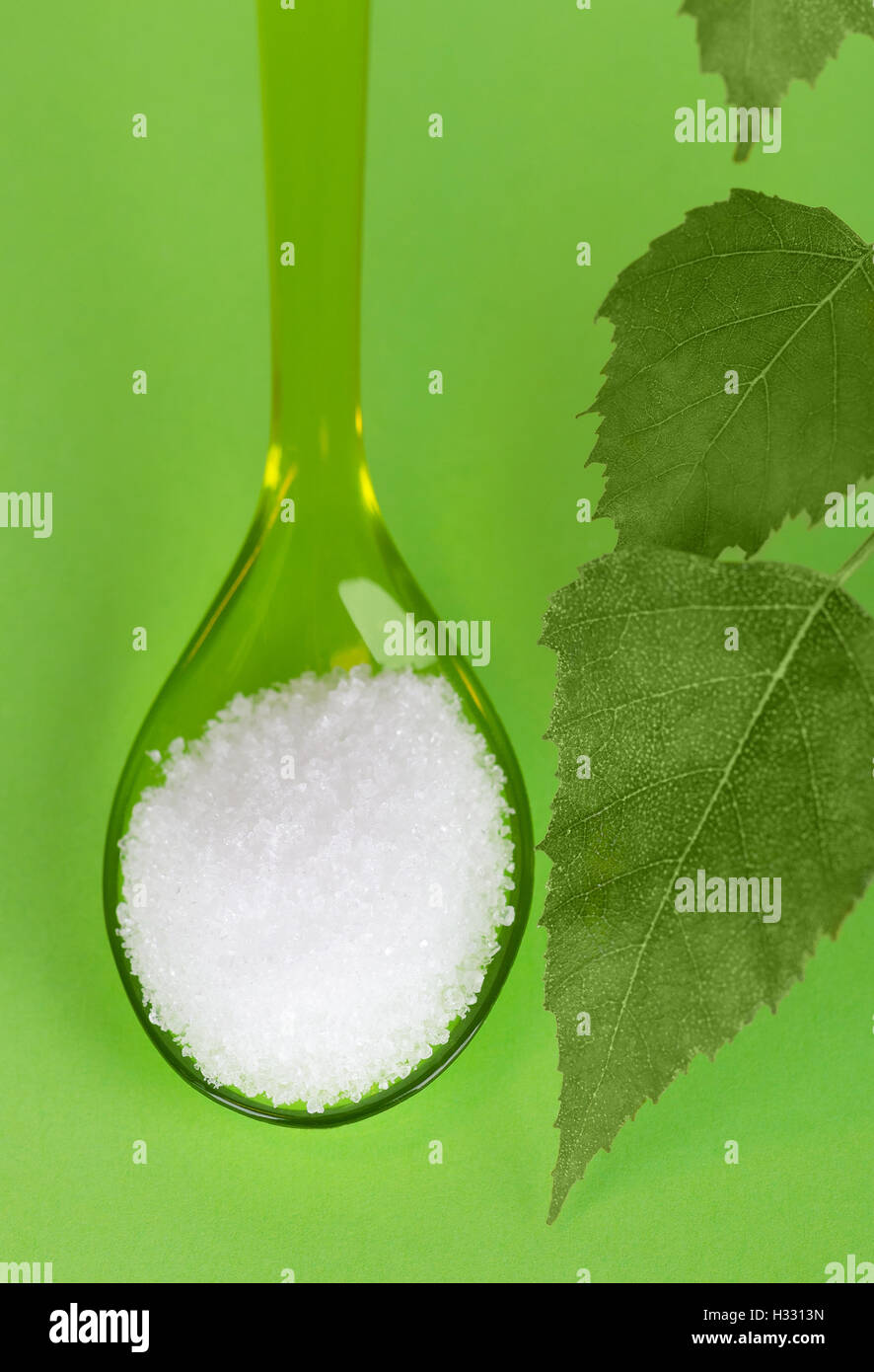 Xylitol birch sugar on plastic spoon with birch leaves over green. White granulated sugar alcohol, substitute used as sweetener. Stock Photo