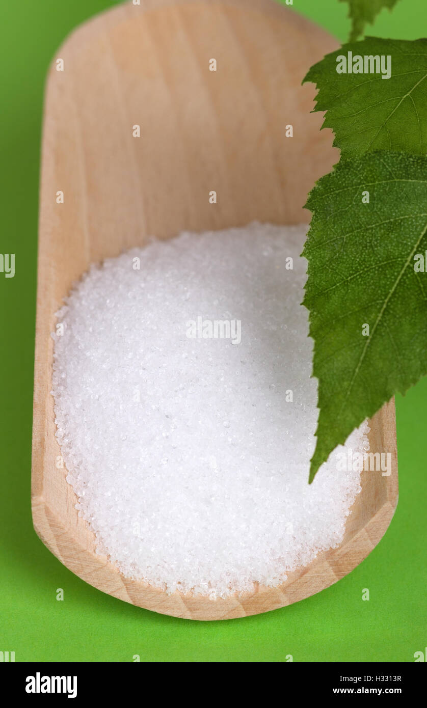 Xylitol birch sugar on wooden scoop with birch leaves over green. White granulated sugar alcohol, substitute used as sweetener. Stock Photo