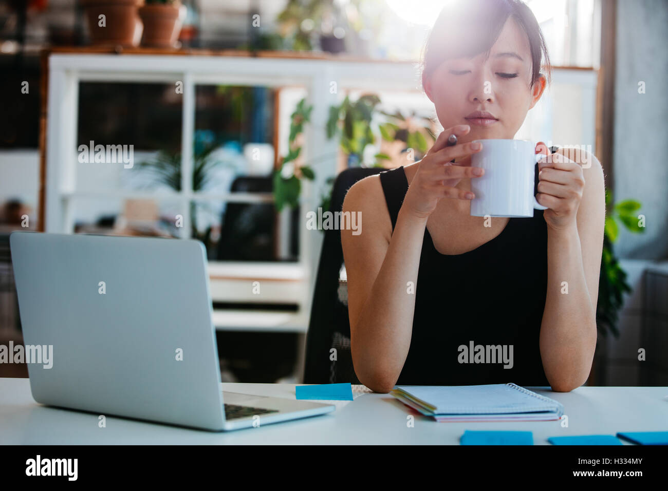 Portrait of relaxed young woman sitting at her desk and having coffee. Asian business woman taking coffee break in office. Stock Photo