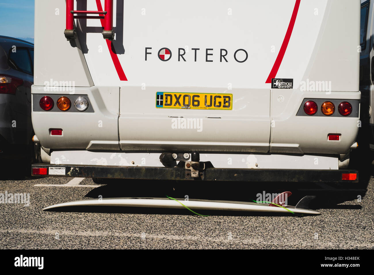 A surfboard laid underneath a Dethleffs Fortero campervan in Sennen Cove car park, Cornwall Stock Photo