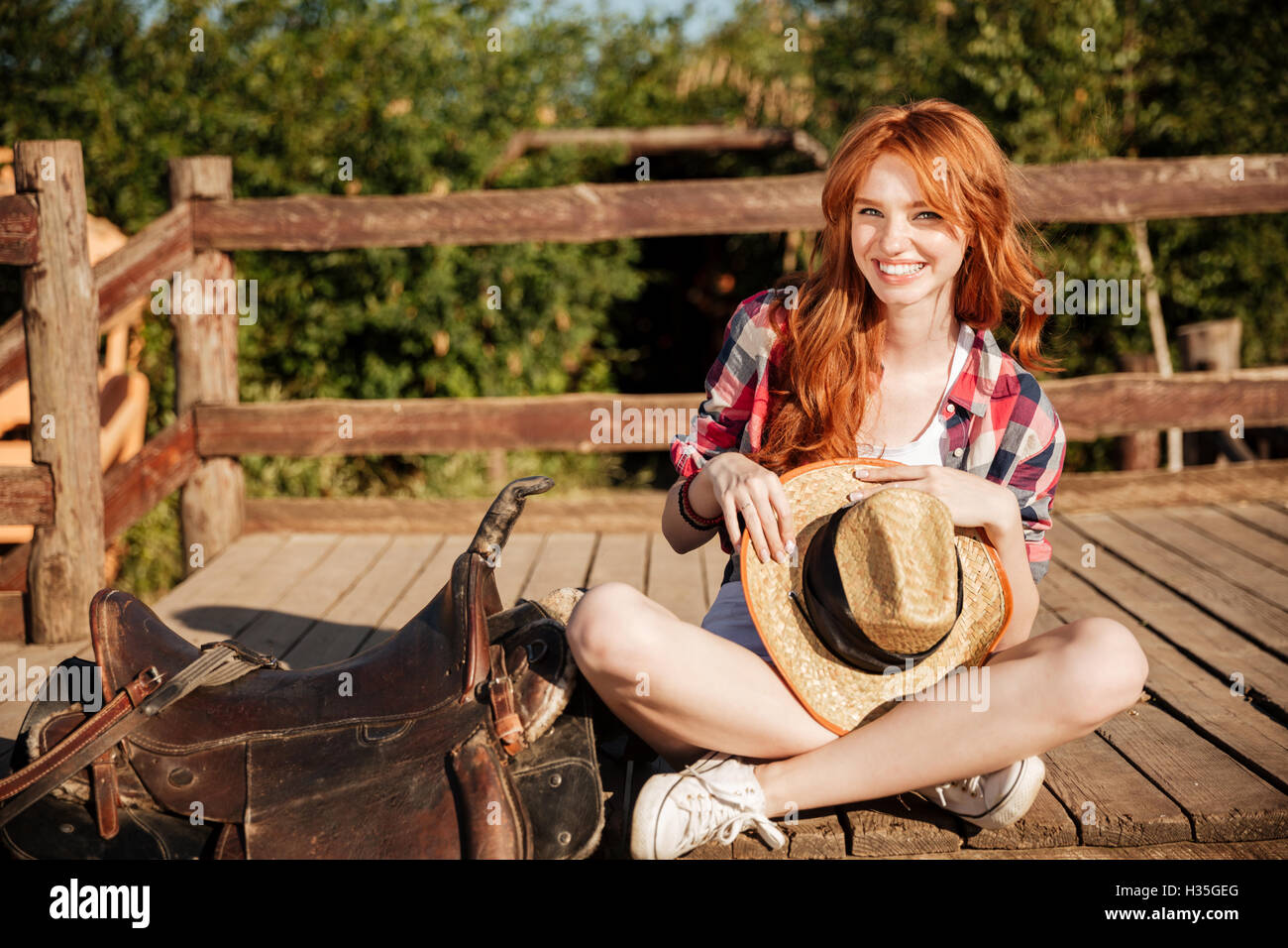 Happy beautiful young woman cowgirl with hat sitting on ranch Stock
