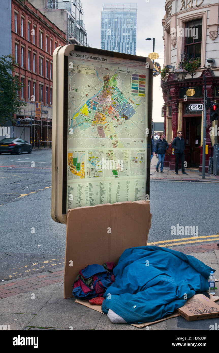 Homelessness; People & passers-by walking past covered homeless beggar, sleeping rough on the pavement against a street map billboard, giving some shelter, on the streets of Manchester, UK. Stock Photo